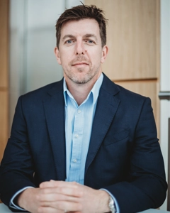 Managing director seated at a table in a navy suit and light blue striped shirt, hands clasped, modern office backdrop